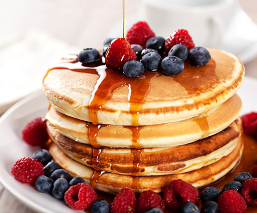 A tower of American style pancakes, with raspberries, blueberries, and maple syrup drizzled over them.