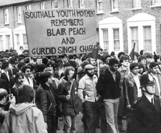 A crowd of people, holding up a banner which is prominent in the photograph. On the banner is written Southall Youth Movement Remembers Blair Peach and Gurdid Singh Chaggab.