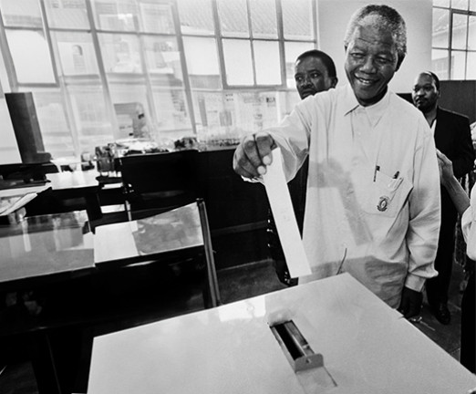 A black and white photograph of Nelson Mandela casting his vote by placing a piece of paper in to a box.