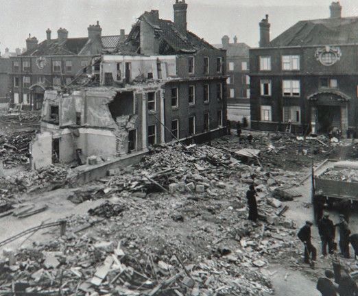 A black and white photograph of rubble in the middle of a street. It shows the destruction of the V-1 Missile. There are shells of houses bordering the street.
