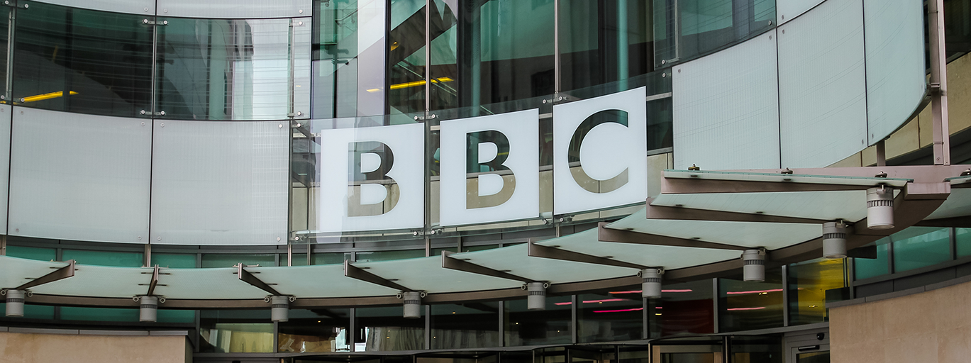 The outside of a BBC building in central London. It is a glass fronted, curved building, with a larger sign which says 'BBC' above the door.