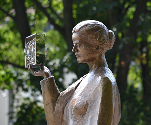 A metal statue of Marie Curie set against a background of green foliage and trees.