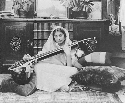 Noor Inayat Khan, a woman with a scarf draped around her head and shoulders, sat cross legged on the floor with a vina, a stringed Indian musical instrument.