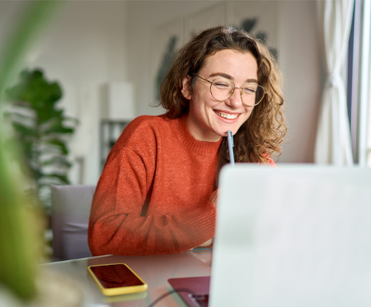 A girl with light brown curly hair, glasses, and an orange jumper smiles as she looks at a laptop screen in the foreground. There is a plant and phone placed on her desk.