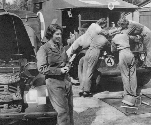 A black and white photograph of the young Princess Elizabeth leaning against a motor vehicle with the bonnet up. She wears overalls and a group of women in the background, in similar dress, can be seen leaning over the open bonnet of another vehicle.