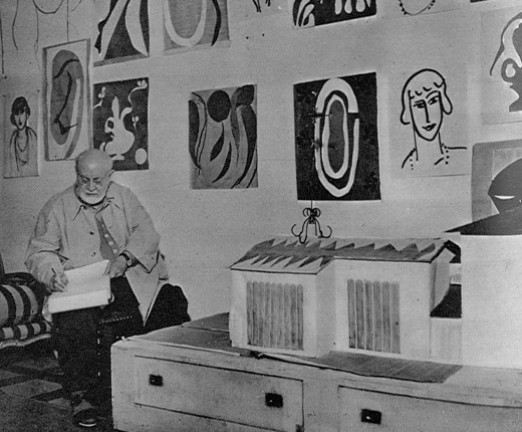 A photograph of Henri Matisse in his studio. There are paintings hanging on the wall behind him.