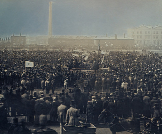 Illustration of a large crowd of people outside some industrial buildings. The crowd faces away, toward the buildings.