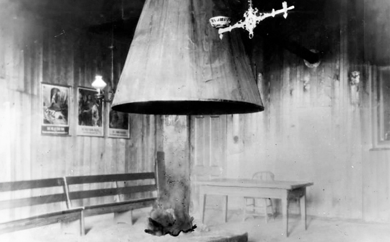 Interior of unidentified mission building, there is a small table and chair in the corner. The photograph is in black and white.