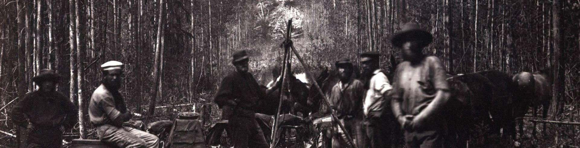 A blurry black and white photograph of a group of people in a forest, stood around a structure of wooden poles