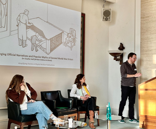 A small lecture hall with a projection of a presentation on the screen. In front of the screen, Charlie Hall stands at a wooden lectern as he presents to the room. Mary Wills sit in a chair beside him, beneath the presentation screen, and beside her sits Beth Potter.