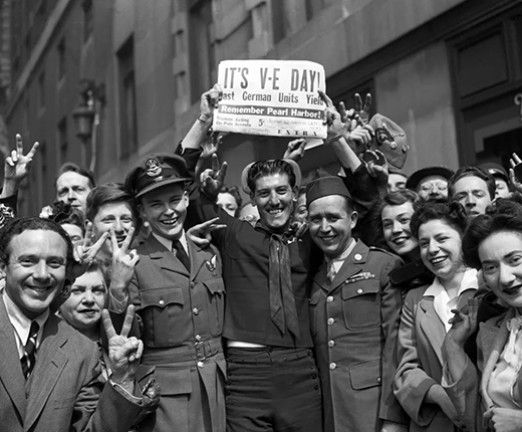 A black and white photograph of people celebrating the end of the Second World War. The man central to the frame holds up a newspaper that reads IT'S V-E DAY. Everyone is smiling.