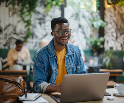 A young man sat at a desk with a laptop in front of him. He wears glasses and is smiling at the screen. Beside his laptop, there is a brown leather bag, and a cup of coffee.
