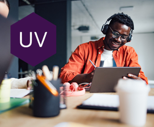 A man wearing headphones looks down at a laptop on the desk before him. He is smiling. There is a purple hexagon overlayed with the letters UV inside it.