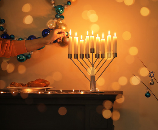 An arm reaches in to frame to light a menorah. There is a mirror hanging on the wall and lots of bokeh lights around with menorah.