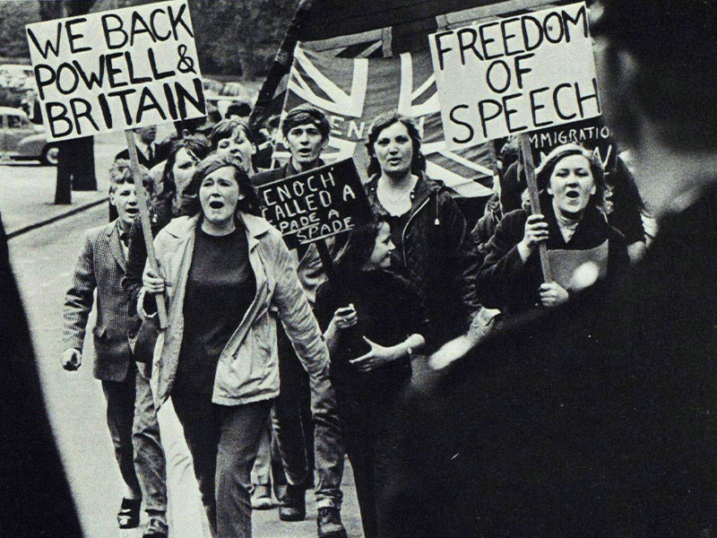 A photograph taken from over the shoulders of two policemen. A crowd of people march towards them holding signs. The two notable signs read We Back Powell & Britain and Freedom of Speech.