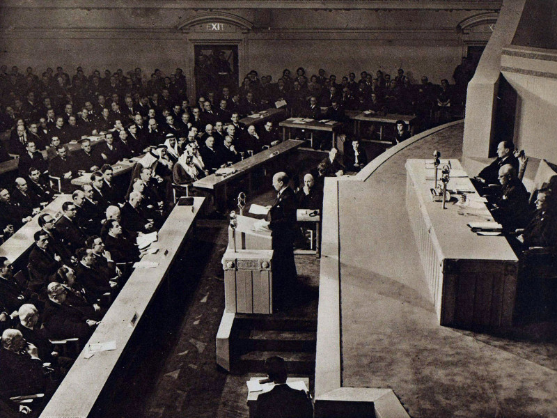 A photograph of the first session of the general assembly of the United Nations Organisation, 10 January 1946. Clement Atlee stands at a podium, addressing a room of people sat at long tables. A table of three men are sat behind him, on a stage.